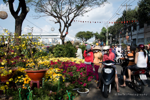 Eine Gruppe von Menschen in Helmen und Mützen, einige gehen und einige fahren Fahrräder auf einer Straße, mit Pflanzen, einem Blumentopf auf einem Hocker, Bäumen, einem Pfahl, Drähten, einer Lampe, einer Brücke und Gebäuden im Hintergrund.