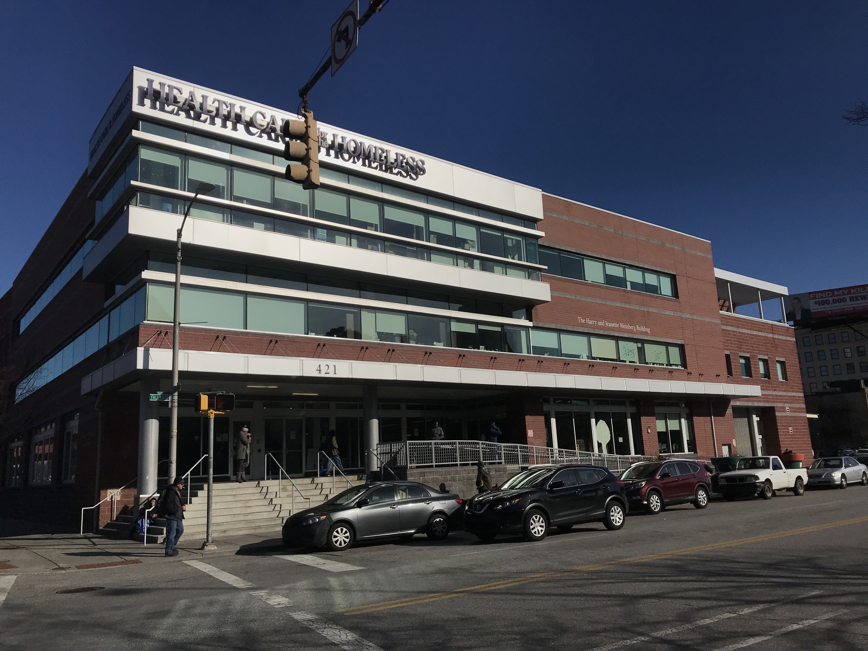 Großes Gebäude mit der Aufschrift 'New York City Health Care Homeless Center' mit parkenden Autos, Fußgängern, Straßenmöblierung und Bäumen unter einem klaren Himmel.