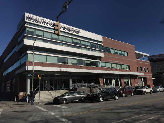 Großes Gebäude mit der Aufschrift 'New York City Health Care Homeless Center' mit parkenden Autos, Fußgängern, Straßenmöblierung und Bäumen unter einem klaren Himmel.