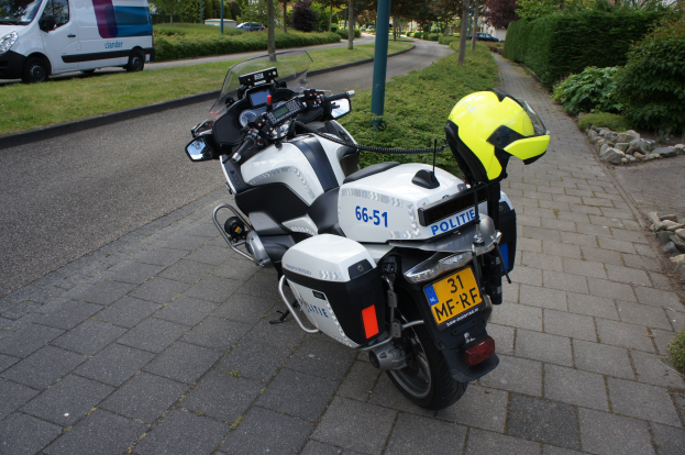 Ein Polizeimotorrad am Straßenrand geparkt mit einem Helm darauf, umgeben von Bäumen, Pfählen, Gebäuden, Pflanzen, Gras, Steinen und anderen Fahrzeugen im Hintergrund.