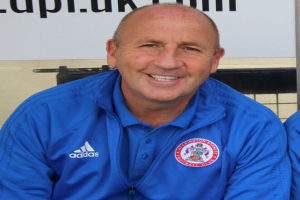 Zwei Männer in einem Raum, einer in einer blauen Jacke, der neben einem anderen Mann sitzt und lächelt, mit einer Wandtafel im Hintergrund.