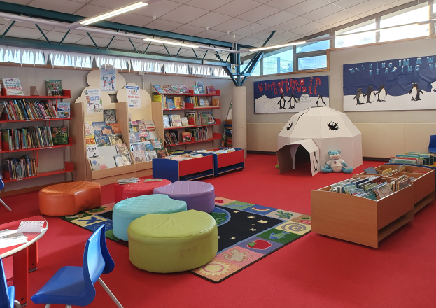 Eine hell erleuchtete Kinderbibliothek mit Bücherregalen, Tischen, Stühlen, Teppichen, Wandtafeln und Deckenleuchten.