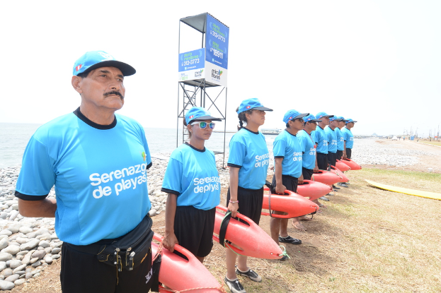 Eine Strandszene mit Menschen, trockenem Gras und Steinen im Vordergrund, einem Surfbrett auf der rechten Seite, einer Leiter und einem Zelt in der Mitte und Schiffen im Wasser unter einem sonnigen Himmel.