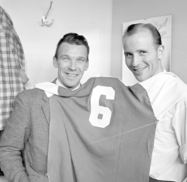 Zwei Männer in Schwarz-Weiß, die strahlend eine Fußball-Trikot hochhalten, mit einer Tafel und einem Bügel an der Wand dahinter.