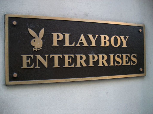 Ein rechteckiges Namensschild mit Logo und Buchstaben, das an einer Wand mit sichtbaren Schrauben befestigt ist.