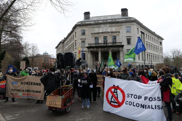 Große Gruppe von Menschen bei einer Demonstration gegen fossile Brennstoffe, die Schilder und Fahnen tragen, mit einem Fahrzeug im Vordergrund und Gebäuden, Bäumen und einem klaren blauen Himmel im Hintergrund.