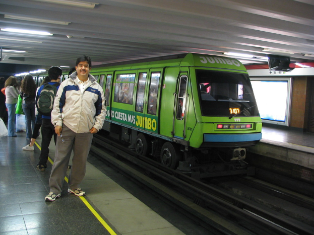 Ein Zug auf Schienen mit Menschen links und einem Dachgestell oben im Bild.