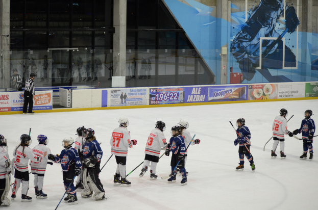 Gruppe junger Menschen, die Eis Hockey auf einer Indoor-Eisfläche spielen, mit Helmen und Stöcken, sowie einer Person links daneben, Bannern, Säulen und Glaswänden im Hintergrund.