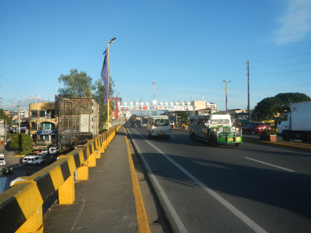 Eine stadtstraßen mit starkem verkehr, eine mittellinie, ein flaggenmast mit einer flagge, gebäude, bäume, elektrischen masten mit drähten und ein bewölkter himmel im hintergrund.