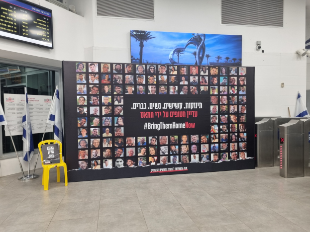 Eine große Wand zeigt mehrere Fotos, einen gelben Stuhl, israelische Regierungsflaggen auf Stangen, eine Tafel mit der Aufschrift "Bring Them Home Now", einen montierten Bildschirm, Deckenleuchten und andere Gegenstände.