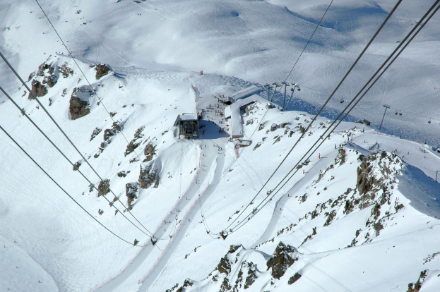 Eine Luftaufnahme eines Skilifts, der einen schneebedeckten Berg hinabfährt, mit Seilbahnseilen, Felsen und Häusern im Hintergrund, bei strahlendem Sonnenschein.