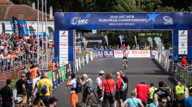 Radfahrer fahren unter einem Bogen mit der Aufschrift "2019 UEC MTB Europameisterschaften" entlang, umgeben von Zuschauern und Bäumen, Gebäuden und einem klaren Himmel im Hintergrund.