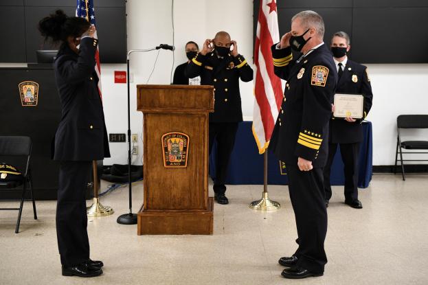Gruppe uniformierter Personen in Masken vor einem Rednerpult mit Mikrofon, umgeben von Fahnen, Stühlen, Tischen und einer Wand während einer Amtseinführung, wobei eine Person ein Zertifikat hält.