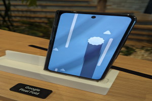 Ein Samsung Galaxy Fold 2 Smartphone mit gebogenem Display und Metallrahmen steht auf einem Tisch mit einer Namensschild, umgeben von Pflanzen und einer Wand im Hintergrund.