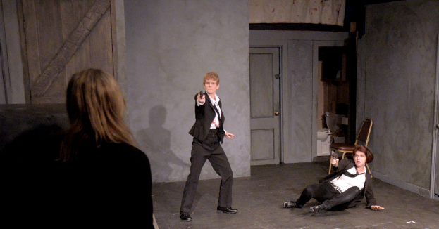 Zwei Frauen in schwarzen Anzügen, eine sitzend auf dem Boden eine Waffe haltend, die andere stehend und eine Waffe haltend, mit einer dritten Frau daneben, in einem Raum mit einem Stuhl, einer Toilette und einer hölzernen Tür.