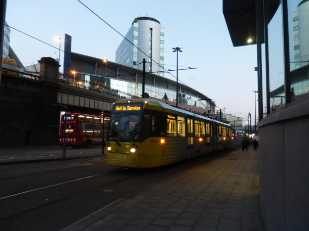 Ein Bus fährt auf einer Straße mit Gebäuden auf beiden Seiten, jedes mit Glasfenstern, und eine Brücke ist auf der linken Seite zu sehen.