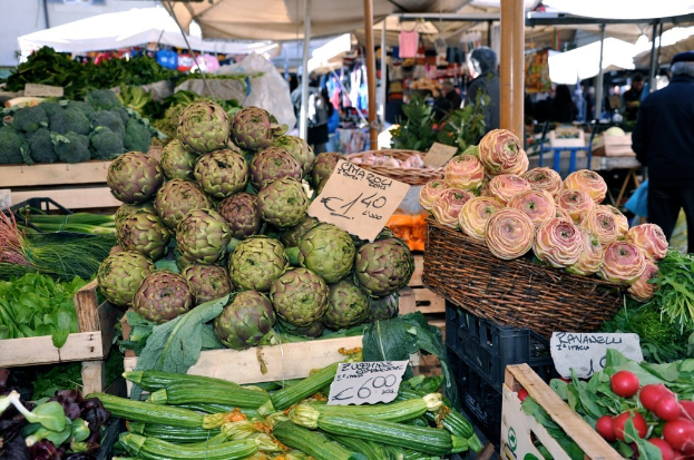 Körbe gefüllt mit Obst, Gemüse und kleinen Tafeln mit Text, mit Zelten und einigen Menschen im Hintergrund.