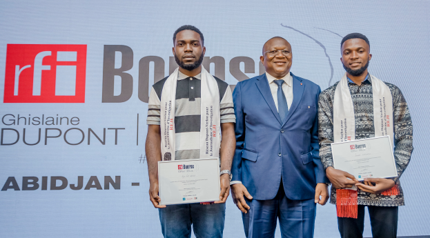 Drei Männer stehen nebeneinander auf einer Bühne, jeder hält ein Zertifikat in der Hand, mit einem Banner im Hintergrund mit der Aufschrift "Ghislaine Dupont Wins RFI Bourse Award."