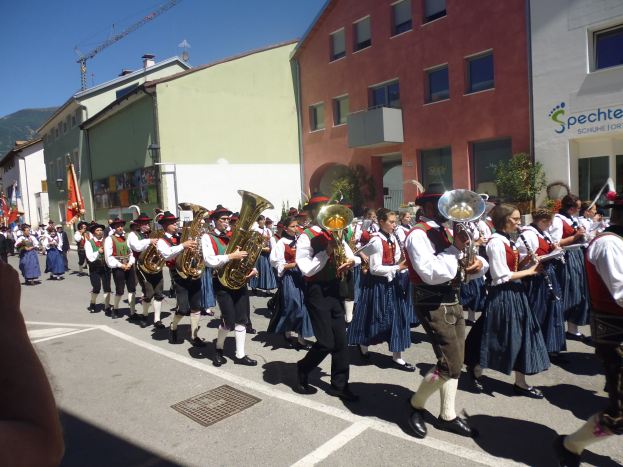 Eine Gruppe von Menschen in traditioneller bayrischer Tracht, die auf der Straße musizieren und marschieren, während sie von Gebäuden gesäumt sind, einige halten Fahnen, mit einem Hügel und einem klaren blauen Himmel im Hintergrund.