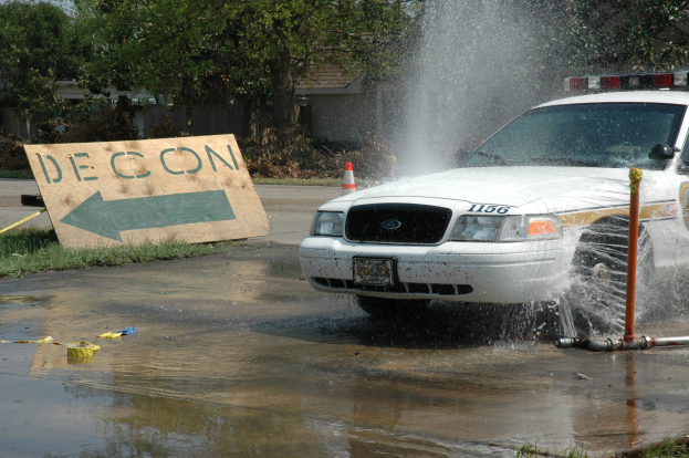 Ein Polizeiwagen fährt durch eine Pfütze, mit einem Schild, einer Verkehrskegel und anderen Gegenständen am Boden, vor einem Hintergrund aus Bäumen, Gebäuden und einem klaren blauen Himmel.