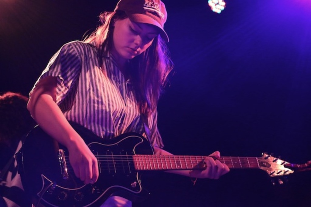 Eine Frau mit einer Mütze spielt eine Gitarre auf der Bühne eines Musikkonzerts, während zahlreiche Lichter den Hintergrund erhellen.