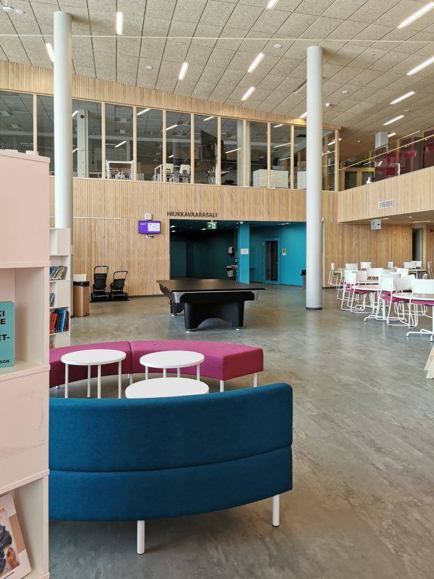 Großer Raum mit mehreren Tischen, Stühlen, Sofas, Hockern, Regalen, Säulen, Deckenleuchten und einem Wandfernseher, ähnlich einem Schulbibliotheksinneren.