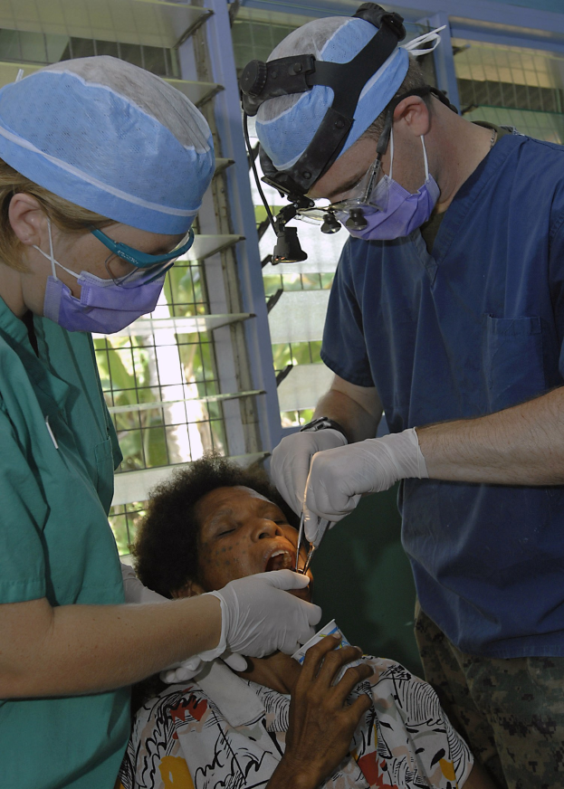 Zwei Chirurgen in Masken und Handschuhen führen eine Gesichtsoperation an einer Frau durch, die auf einem Bett liegt, mit Bäumen im Hintergrund durch ein Fenster sichtbar.