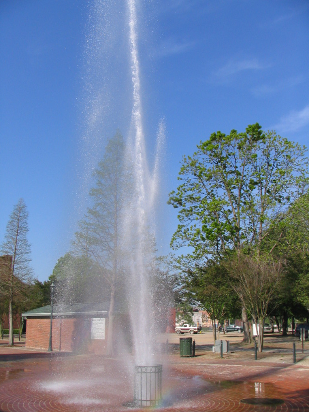 Ein Brunnen in der Mitte, mit einem Haus und Bäumen links, einer kleinen Trommel in der Nähe eines Pfahls rechts und parkenden Fahrzeugen im Hintergrund unter einem Himmel.