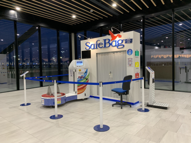 Sicherheitskontrolle für Gepäck am Flughafen mit einem Stuhl, Absperrpollern, Maschinen, einem Bildschirm, einer bedruckten Tafel, Glaswänden, Deckenleuchten und sichtbarem Boden.