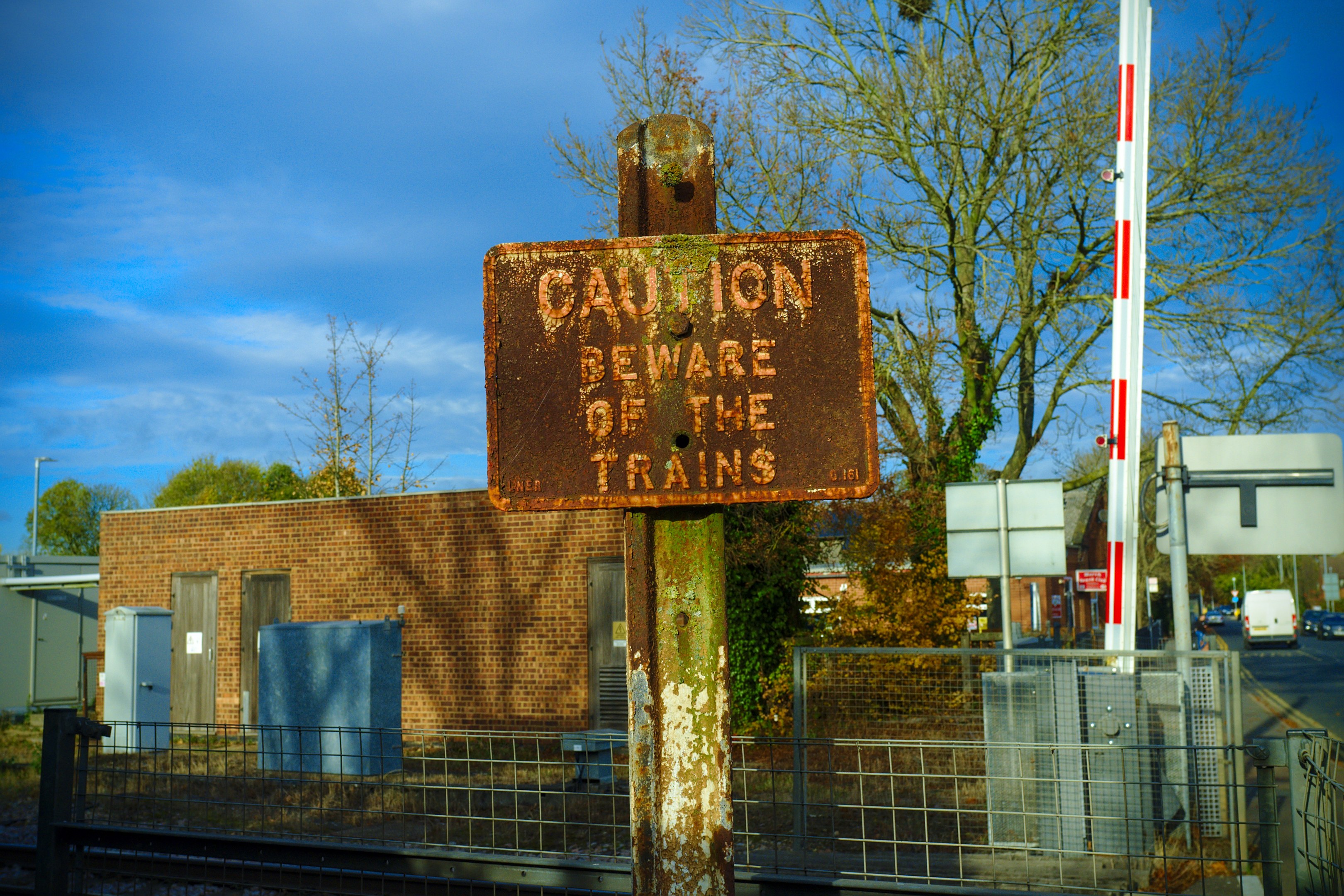Vorsichtsschild auf einem Bahngleis umgeben von einem Metallzaun, Bäumen, Strommasten, einem Gebäude mit Fenstern, Containern, Fahrzeugen auf einer Straße und einem bewölkten Himmel.