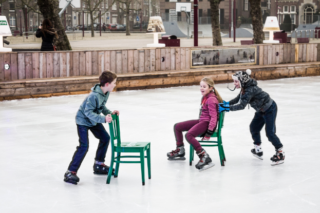 Kinder beim Skifahren vor einem Spielplatz, mit drei Kindern und zwei Stühlen in der Mitte und Gebäuden, Bäumen, Bänken, Pfählen und einem Basketballfeld im Hintergrund.