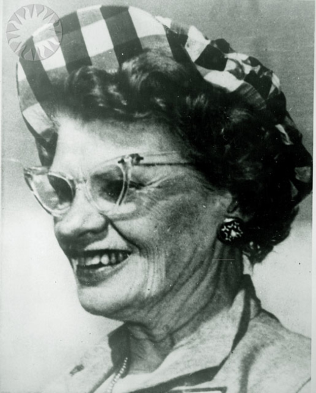 Eine lachende Frau mit Brille und weiß-schwarzem Hut.