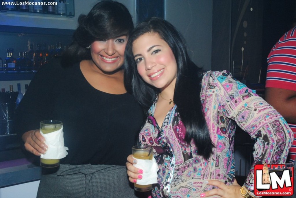 Zwei Frauen mit Gläsern und Taschentüchern, mit Regalen voller Flaschen im Hintergrund und sichtbaren Wasserzeichen auf dem Bild.