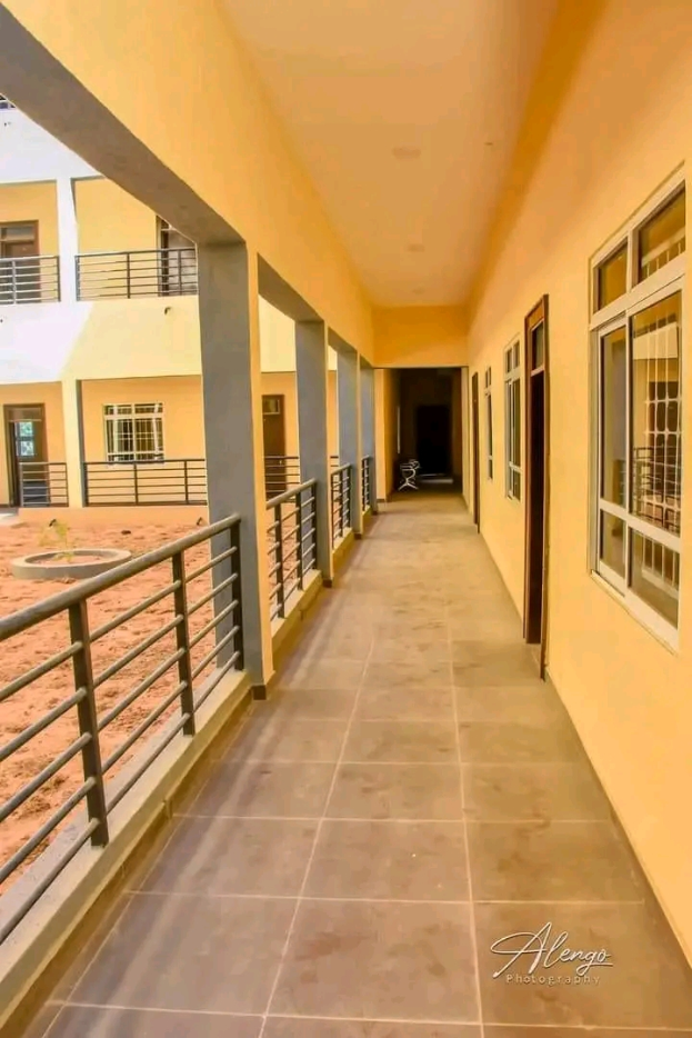 Ein Gebäude mit Fenstern, Türen, Säulen, Geländern und einem langen Flur mit einem gefliesten Boden und einem Balkon, mit anderen Gebäuden und einem klaren blauen Himmel im Hintergrund und Text unten, der "3-Zimmer-Wohnung zur Miete in Ntinda, Kampala" liest.