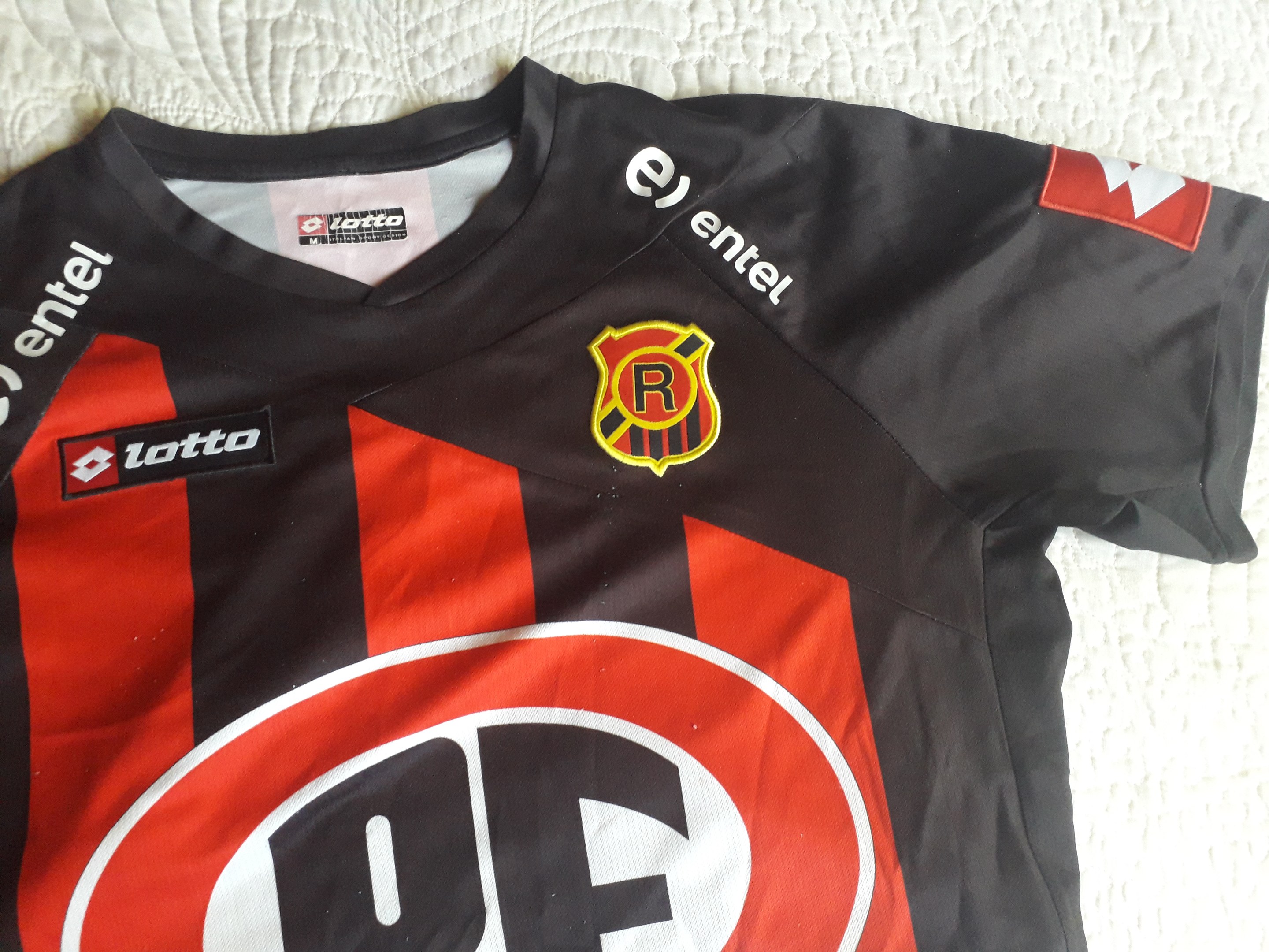 Schwarzer und roter Fußballjersey mit einem roten und schwarzen Streifen, der ein Logo und Text trägt, auf einer weißen Oberfläche angezeigt.