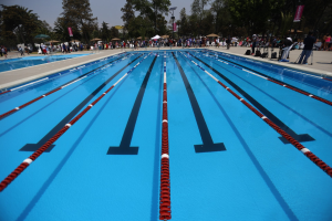 Ein großes Schwimmbad mit Bahnentrennern, umgeben von Menschen mit Taschen und Gegenständen, Zelten, Schildern, Stangen und einem bewölkten Himmel im Hintergrund.