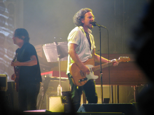 Ein Mann in einem weißen Hemd spielt Gitarre und singt in ein Mikrofon auf der Bühne, neben ihm spielt ein anderer Mann in einem schwarzen Hemd Gitarre; im Hintergrund sind ein Piano, Kabel, Lautsprecher und Lichter zu sehen.