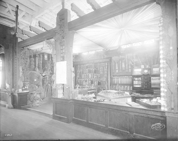 Ein altes Schwarz-Weiß-Foto einer Apotheke mit einer Theke, auf der verschiedene Gegenstände stehen und Regalen im Hintergrund, die mit Flaschen und anderen Gegenständen gefüllt sind.