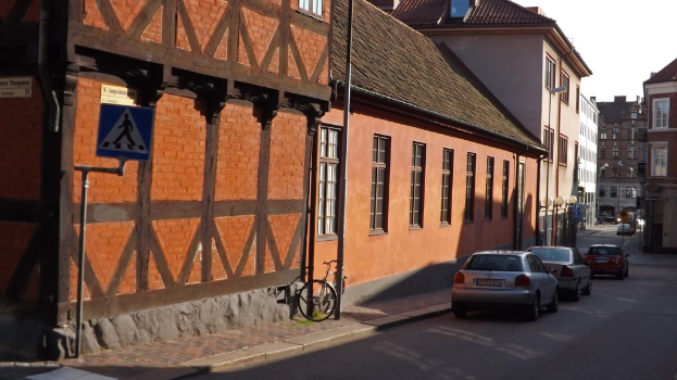 Fahrzeuge auf der Straße mit Gebäuden, Laternen, Schildermasten, Fenstern und Dächern im Hintergrund und einem Fahrrad auf dem Gehweg in der Nähe eines Pfahls.
