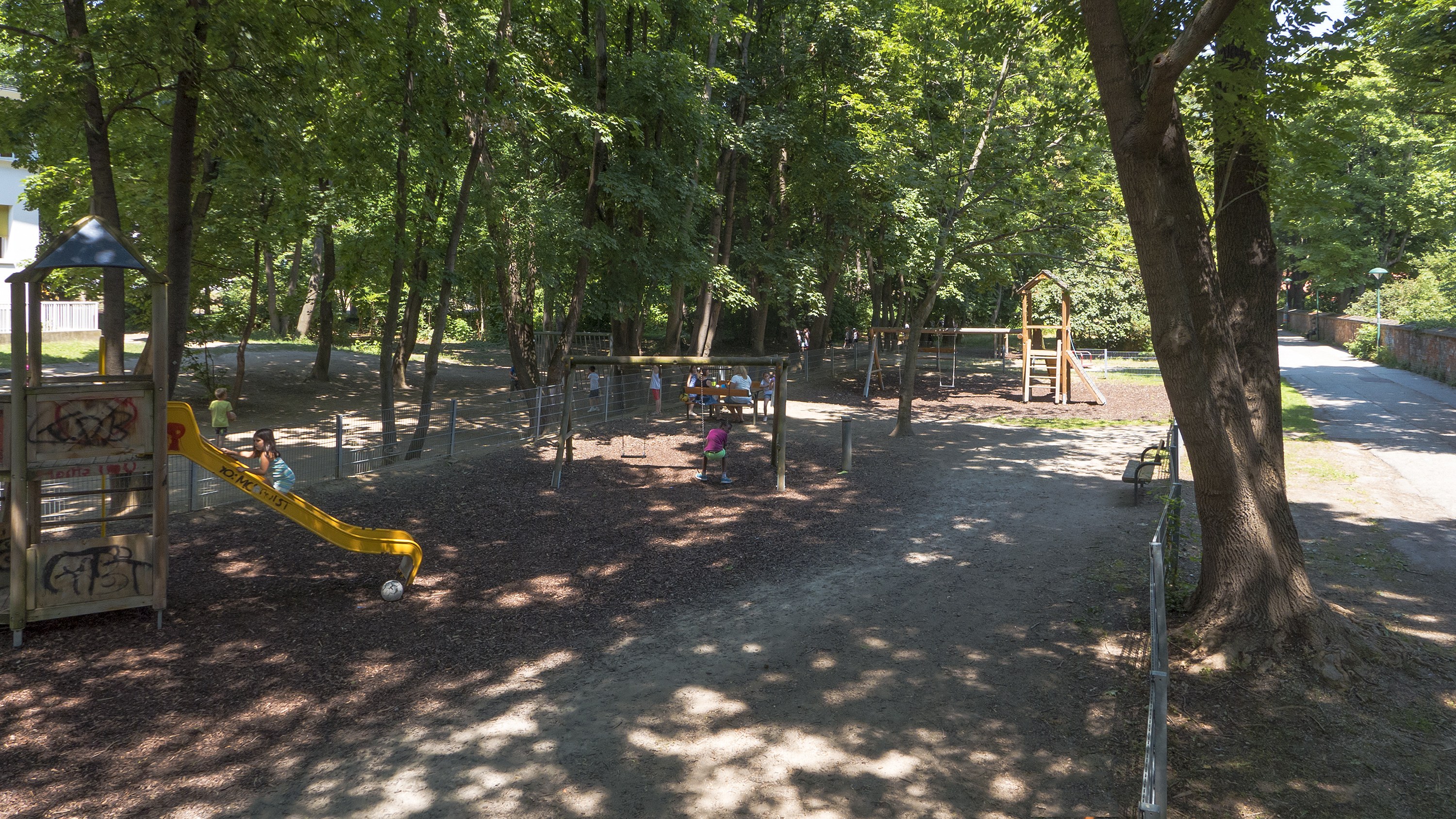 Ein Park mit einem Spielplatz in der Mitte, umgeben von Bäumen, mit ein paar Menschen auf der Ausrüstung und einem Gebäude links daneben.