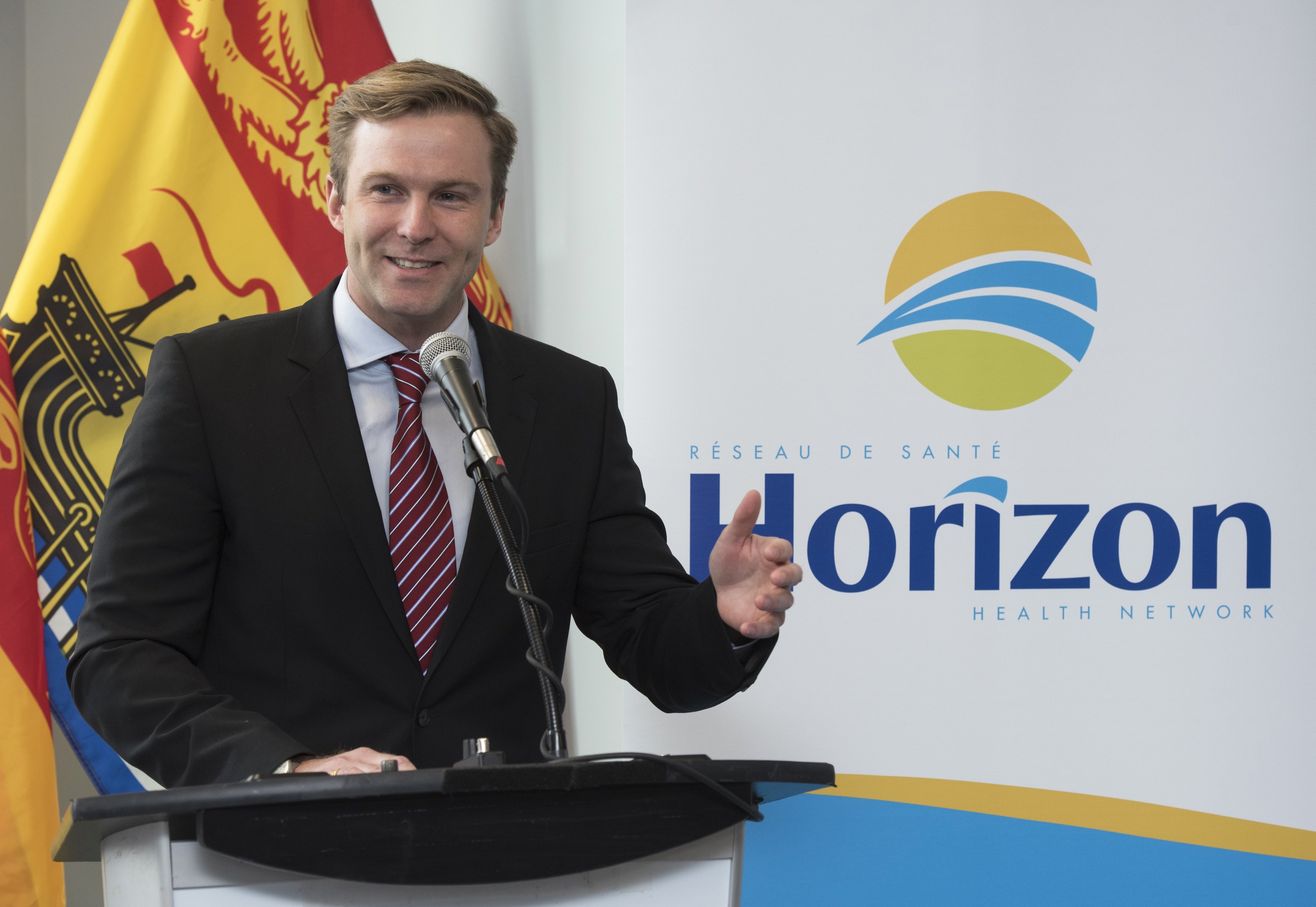 Ein Mann mit einem schwarzen Blazer, weißem Hemd und roter Krawatte steht an einem Podium mit einem Mikrofon und einem Banner im Hintergrund und lächelt.