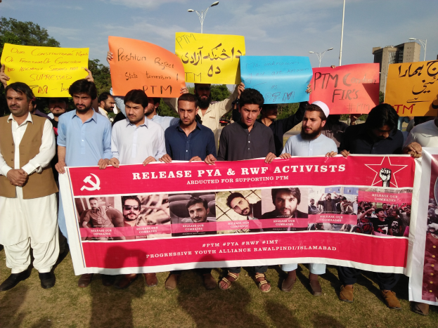 Eine Gruppe von Menschen steht auf Gras, hält eine Tafel mit der Aufschrift "Pya & RWF-Aktivisten", umgeben von Straßenlaternen, Straßenlaternen, Bäumen, Gebäuden und einem bewölkten Himmel, protestiert gegen die Freilassung von Pya- und RWF-Aktivisten.