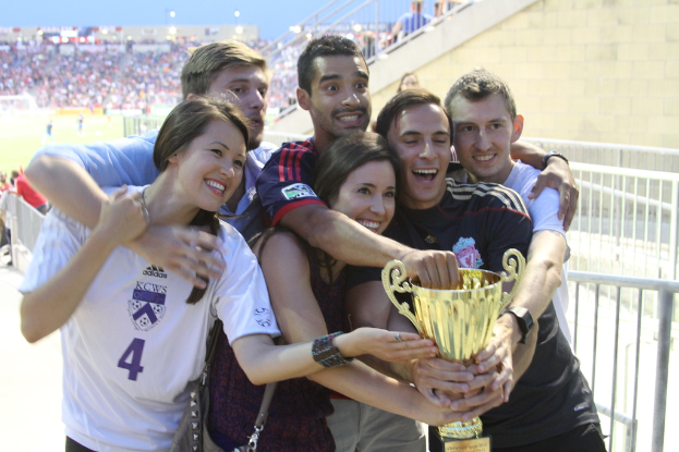 Sechs Personen, zwei Frauen und vier Männer, halten einen Pokal und lächeln, während sie für ein Foto vor einem Eisenzaun posieren, mit einer Wand und einem Zaun oben rechts, Menschen im Hintergrund und sichtbaren Lichtern, möglicherweise in einem Stadion.