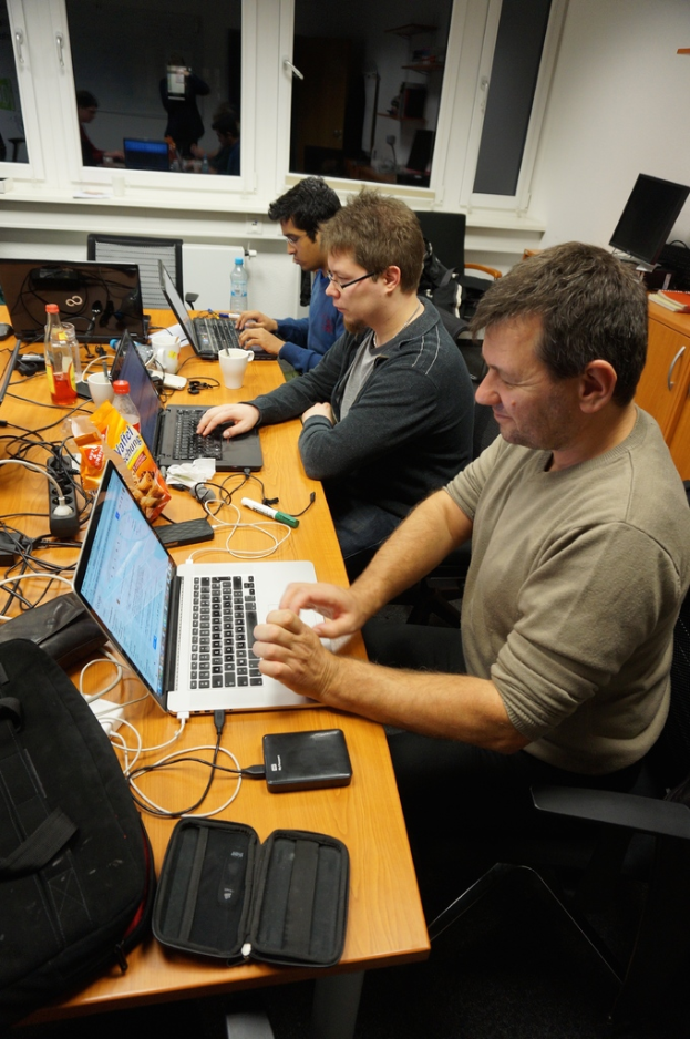 Drei Personen sitzen auf Stühlen und arbeiten an Laptops, mit elektrischen Geräten und einer Flasche und einem Becher auf dem Tisch vor ihnen.
