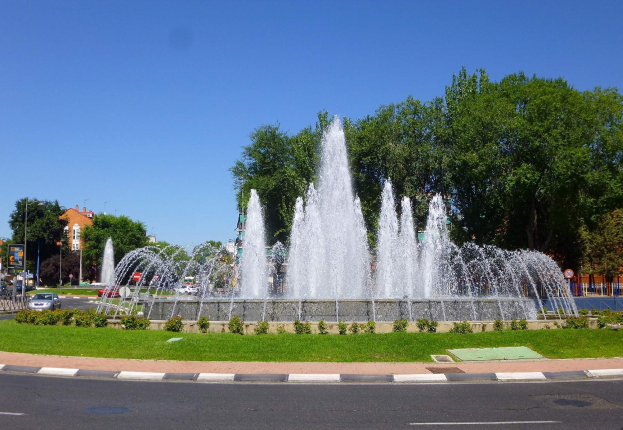 Großer Brunnen auf einem Stadtplatz mit Grünflächen, Fahrzeugen, Laternen, Texttafeln, Gebäuden und einem klaren blauen Himmel.