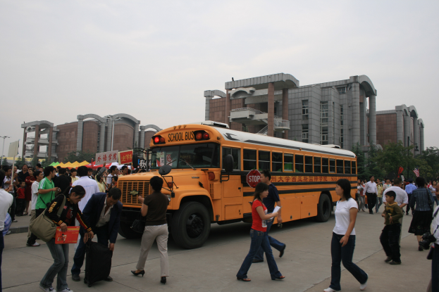 Eine Gruppe von Menschen, einige mit Taschen, geht auf einer Straße vor einem Schulbus, umgeben von Gebäuden, Bäumen und Laternen unter einem klaren blauen Himmel.