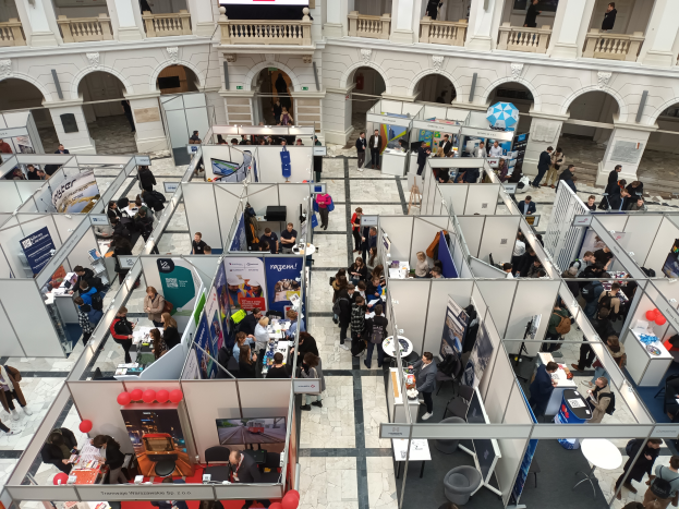 Eine große Gruppe von Menschen versammelte sich in einem Raum voller Stände, Banner, Tische, Stühle und Luftballons, mit einem Gebäude im Hintergrund mit Bögen, Säulen, Geländern und einem Bildschirm.