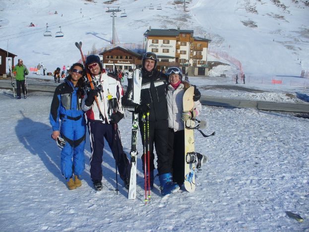 Vier Personen posieren für ein Foto mit Schlittschuhschützern und Werkzeugen, mit Gebäuden und anderen Menschen im Hintergrund, vor einer schneebedeckten Berglandschaft.