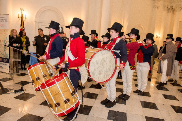 Eine Gruppe von Musikern, die auf einer Bühne in einem Saal mit strukturierten Wänden Trommeln und Flöten spielen, während Menschen um sie herum stehen, mit einem Podium neben der Bühne.