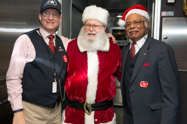 Eine Gruppe von Menschen mit Brillen, Mützen, Krawatten, ID-Karten und Abzeichen, wobei eine Person ein Weihnachtskostüm trägt, vor einer Stahlwand mit Lichtern, Schildern und anderen Gegenständen im Hintergrund steht.